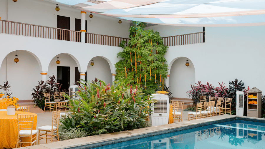 A swimming pool sits next to a courtyard garden, seating area, and white multi-storey building at Bougainvillea, Alibaug.