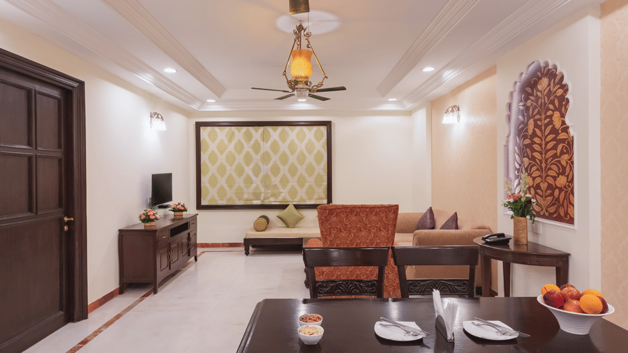 A well lit spacious premier suite dining area at Shri Vallabh Vilas Lords Plaza Nathdwara, featuring a dining set, a living space at the back, wall arts, wooden furnishings, and a T.V.