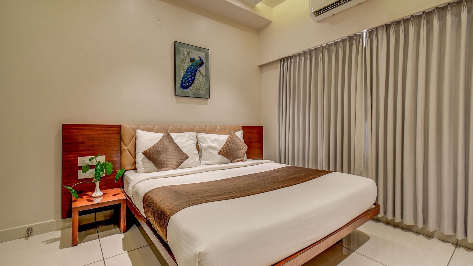 A spacious room at Harigovinda's JNStay with a large bed, a bedside table, large windows fully covered with curtains, and a wall-mounted photo frame above the headboard