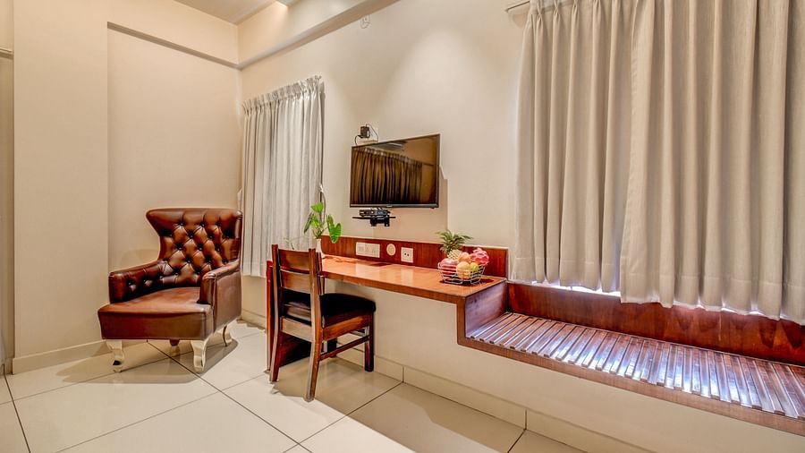 A wall-mounted TV at Harigovinda's JNStay, with a wall-mounted table below it, a cushioned chair beside, and curtains covering the window