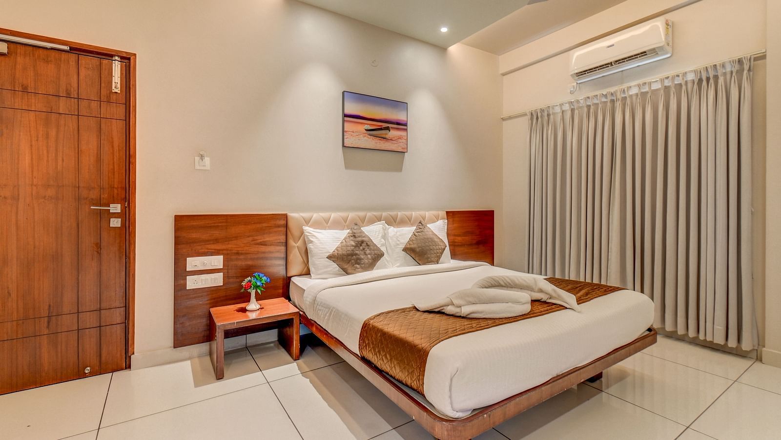 A spacious room at Harigovinda's JNStay with a large bed, wooden headboard, bedside table with a plant, a door, and curtains covering the window