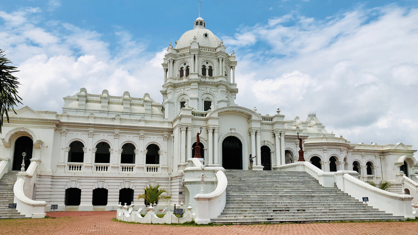 Ujjayanta Palace in Agartala featuring white domed Indo-Saracenic architecture with grand staircase, arched verandas and manicured grounds under blue sky.