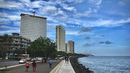 An image of a street of Mumbai near the Marine Drive with high rise buildings where are people are seen walking with a blue sky in the background