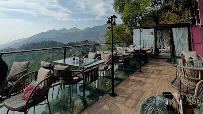A vibrant outdoor cafe balcony at 19th Century A unit of Royal Arc Hospitality featuring tables and chairs, offering panoramic views of a lush valley and distant mountains under a cloudy sky.