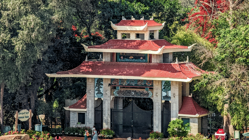 A traditional pavilion-style structure set within a lush garden, framed by tall trees and flowering plants, with a modern high-rise building visible in the background