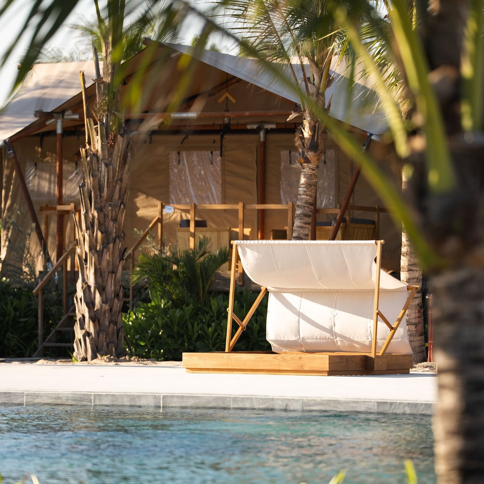 A serene view of the Grateful Tent at WoCAL Retreat Ke Ga, partially framed by palm trees, with a hammock-style lounger placed beside a calm swimming pool. The tent structure in the background features a raised wooden deck and canvas covering, blending nat