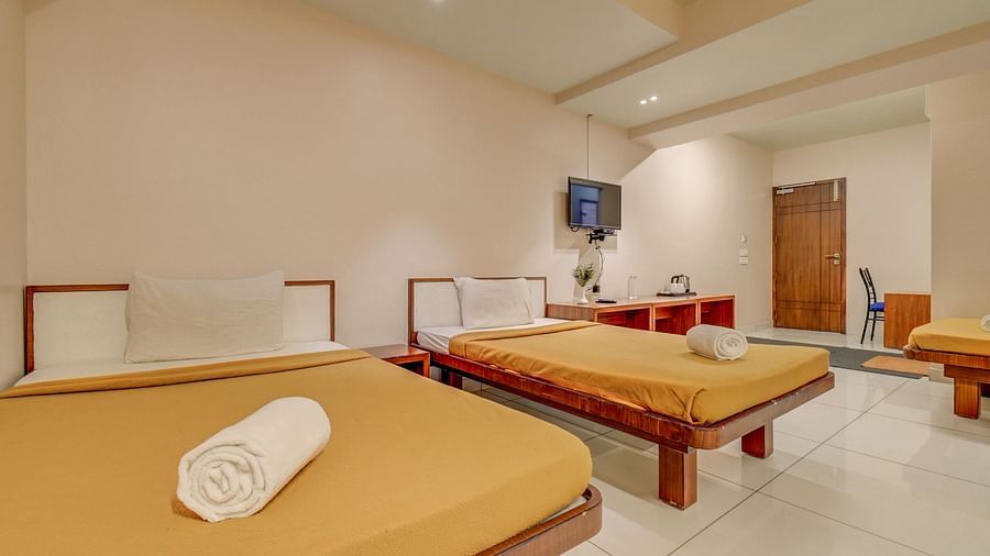A spacious room at Hari Govinda’s JN Stay, Shakthinagar featuring neatly made single beds, a wall-mounted television, and a door at the furthest end