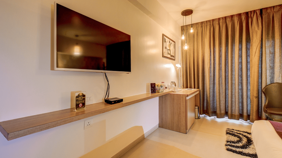 A television screen hangs mounted on a shelf structure opposite a bed frame near curtains. This perspective illustrates the entertainment viewing area from within a Suite Room at The SSK Solitaire Hotel & Banquets, Mumbai Naka - Nashik.
