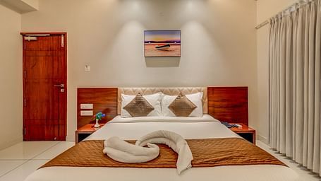 A spacious bed at Harigovinda's JNStay with neatly made bedding, towels arranged in a decorative pattern, a wall-mounted photo frame above, and a door to the side
