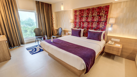 A bed faces a large window while a chair and small table sit in the corner of the floor area. This view shows the bedroom setup of a Suite Room at The SSK Solitaire Hotel & Banquets, Mumbai Naka - Nashik.