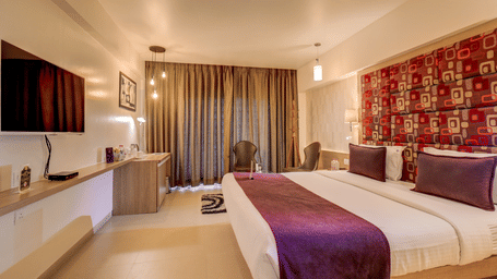 A bed sits positioned against a patterned headboard wall next to a television screen mounted on a ledge. This layout captures the sleeping quarters of a Suite Room at The SSK Solitaire Hotel & Banquets, Mumbai Naka - Nashik.