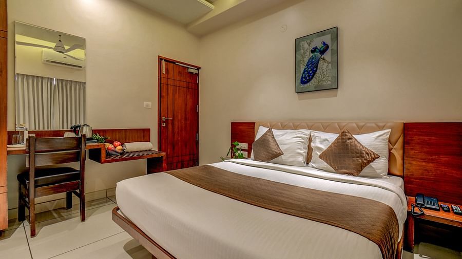 A spacious room at Harigovinda's JNStay with a large bed, wooden headboard, bedside table, photo frame above, and a side table with a wall-mounted mirror and chair