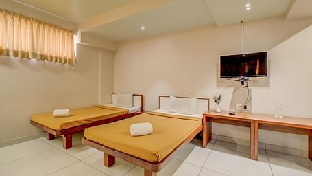 A spacious room at Harigovinda's JNStay, featuring a wall-mounted TV above a wooden table set against the wall, and two beds