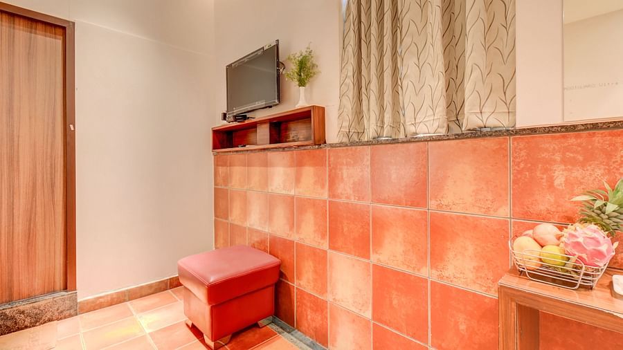 A wall-mounted TV, a cushioned chair, a window covered with curtains, and a table with a fruit basket at Harigovinda's JNStay