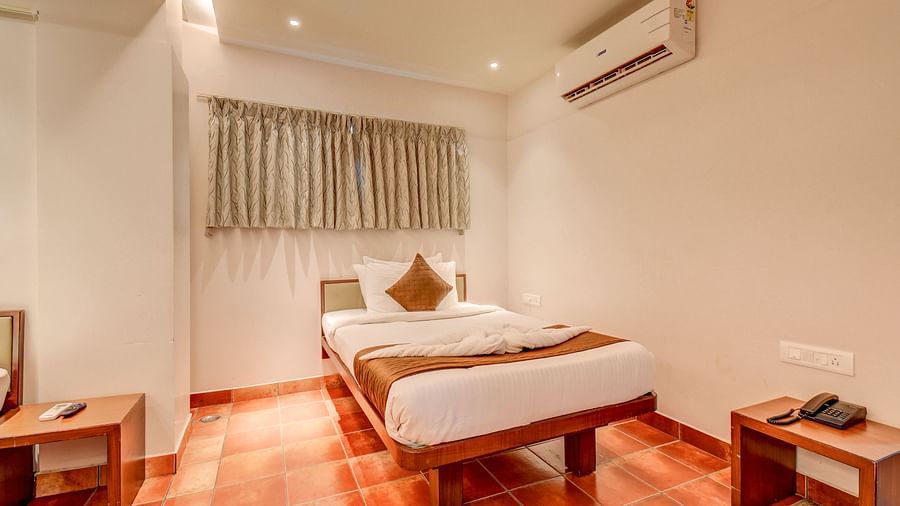 A spacious room at Harigovinda's JNStay with a bed, two wooden side tables, a wall-mounted AC, and high windows covered with curtains