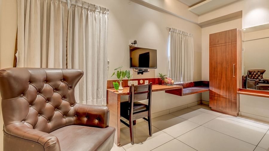 A spacious room at Harigovinda's JNStay with a cushioned chair, wall-mounted TV, desk below it, and a chair