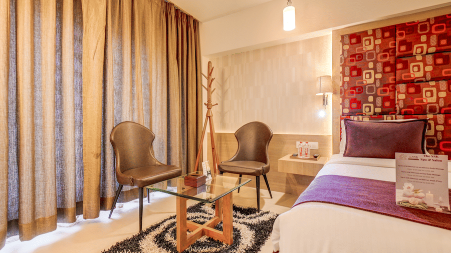 Two chairs and a table are placed on a rug near a bed and a window covered by curtains. This seating arrangement offers a relaxation spot within a Suite Room at The SSK Solitaire Hotel & Banquets, Mumbai Naka - Nashik.
