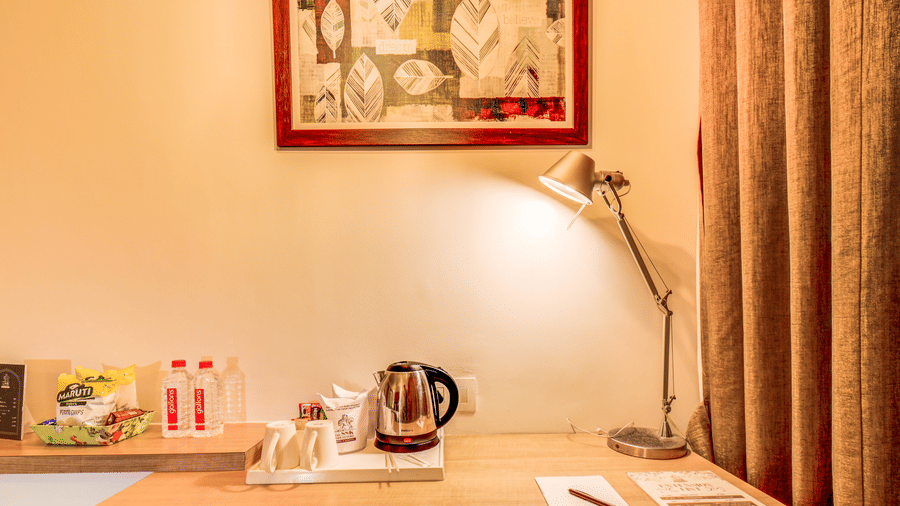 A kettle, bottles, and stationery sit arranged on a wooden desk surface beneath a lamp and a framed picture. This image displays the work desk setup in a Suite Room at The SSK Solitaire Hotel & Banquets, Mumbai Naka - Nashik.