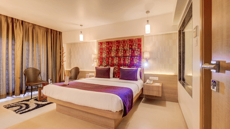 A bed sits positioned between a window with curtains and a wall opening near a doorway. This view shows the layout of the sleeping area in a Suite Room at The SSK Solitaire Hotel & Banquets, Mumbai Naka - Nashik.