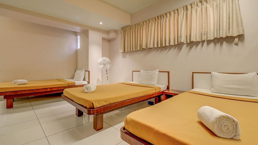 A spacious room with three neatly made beds, each with rolled towels placed over the blanket, and a rectangular window panel covered with curtains at Harigovinda's JNStay