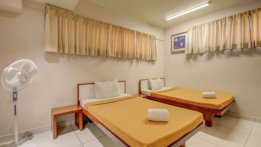 A spacious room at Harigovinda's JNStay, featuring two beds, a bedside table, two adjustable window panels covered with curtains, and a ceiling fan on the side