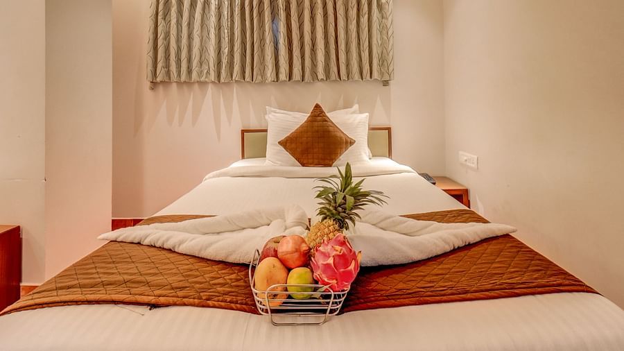 A spacious bed at Harigovinda's JNStay, with neatly arranged blankets, pillows, and a fruit basket, with windows covered by curtains above