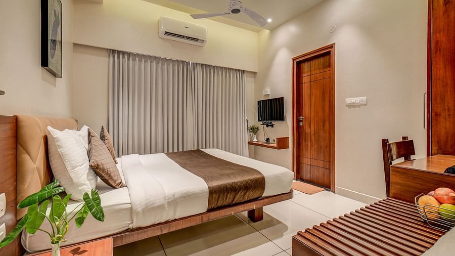 A spacious room at Harigovinda's JNStay with a bed, bedside table with a small plant, a table with fruits, and curtains covering one side of the wall