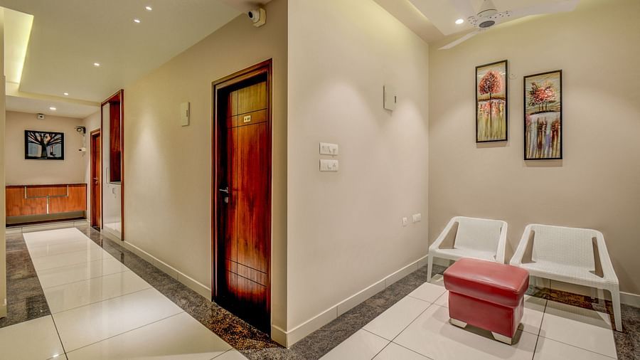 Corridor view at Harigovinda's JNStay, with one side leading to rooms and the other to a sitting area with two chairs and a small cushioned table below wall-mounted photo frames