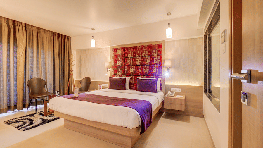 A bed sits positioned between a window with curtains and a wall opening near a doorway. This view shows the layout of the sleeping area in a Suite Room at The SSK Solitaire Hotel & Banquets, Mumbai Naka - Nashik.