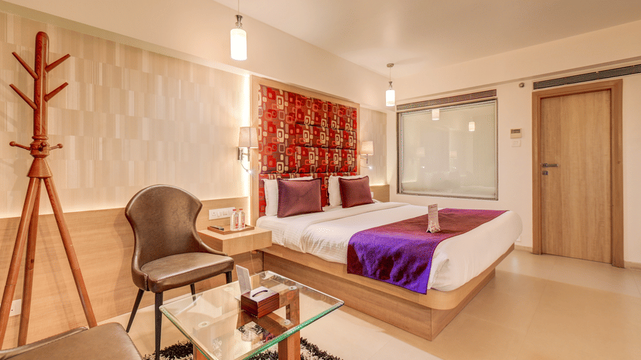 A coat stand and chairs sit near a bed positioned against a patterned headboard wall. This layout shows a perspective of the interior space inside a Suite Room at The SSK Solitaire Hotel & Banquets, Mumbai Naka - Nashik.