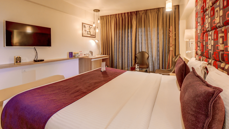 A bed with pillows stands positioned opposite a wall structure holding a television monitor. This wide angle shows the sleeping quarters and media setup inside a Suite Room at The SSK Solitaire Hotel & Banquets, Mumbai Naka - Nashik.