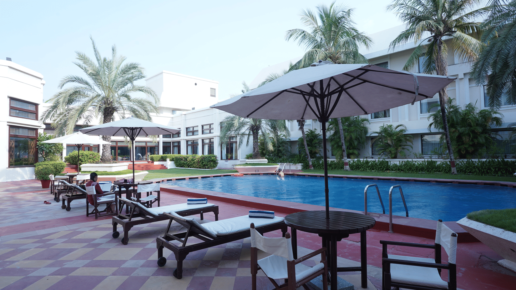 Outdoor swimming pool at The Ummed Ahmedabad with lounge chairs and relaxing resort-style ambiance surrounded by palm trees.