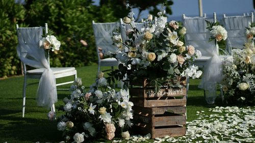 Rose petals cover green grass in a sunny outdoor wedding setting with chairs draped in fabric and large floral arrangements in wooden crates.