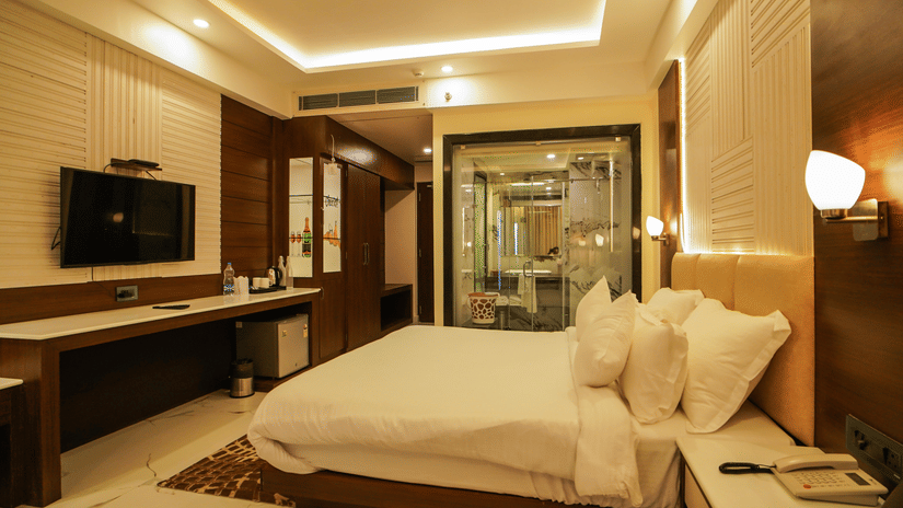 A wall-mounted television, desk, and a well-made bed face a transparent glass bathroom inside a bedroom at Shanti Seaview Resort & Spa.