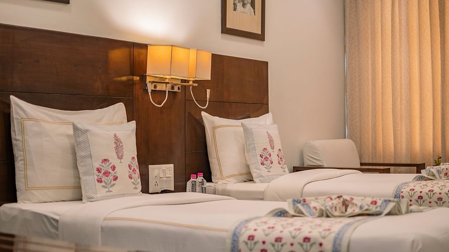 Twin beds with floral cushions and wall-mounted lights at Clarks Collection Vadodara, featuring bottled water and framed photos above.