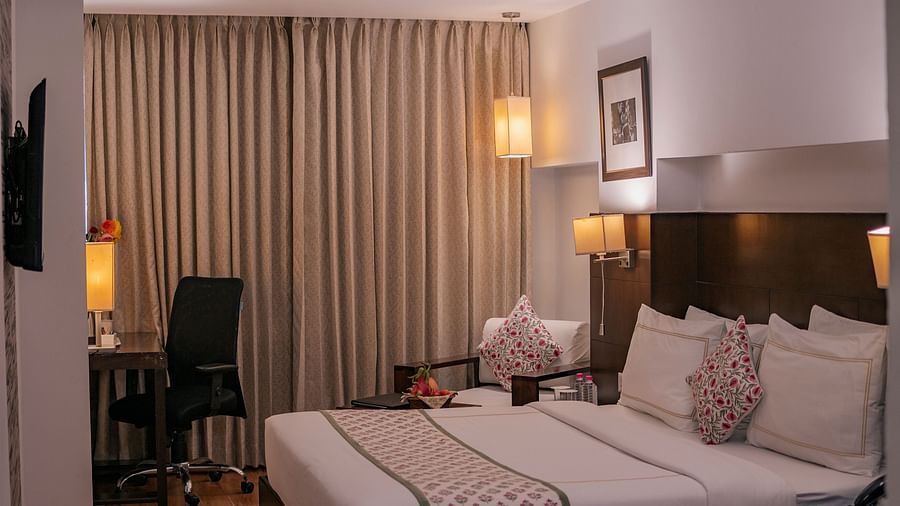 Wide view of twin beds, wall-mounted TV, desk with kettle and flower vase in a room at Clarks Collection Vadodara.