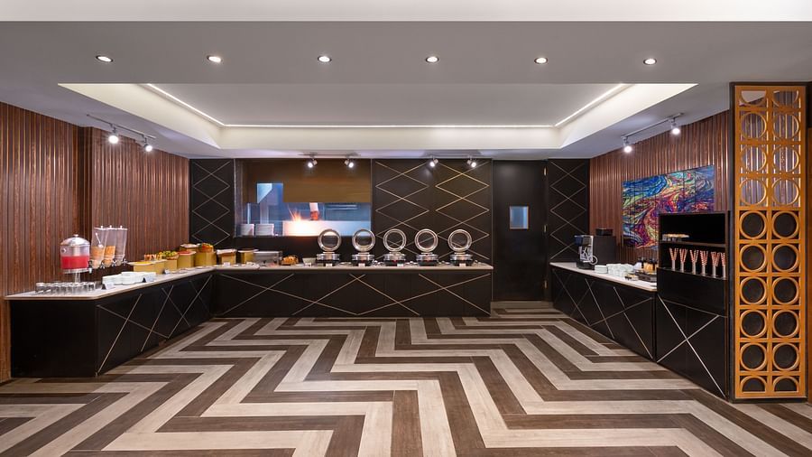 A spacious Hall with a L shaped counter and various cooking utensils on them to keep the food warm along with a juice dispenser and zigzag patterns on the floor - Clarks Inn Express, Near Jammu Bari Brahmana Railway Station