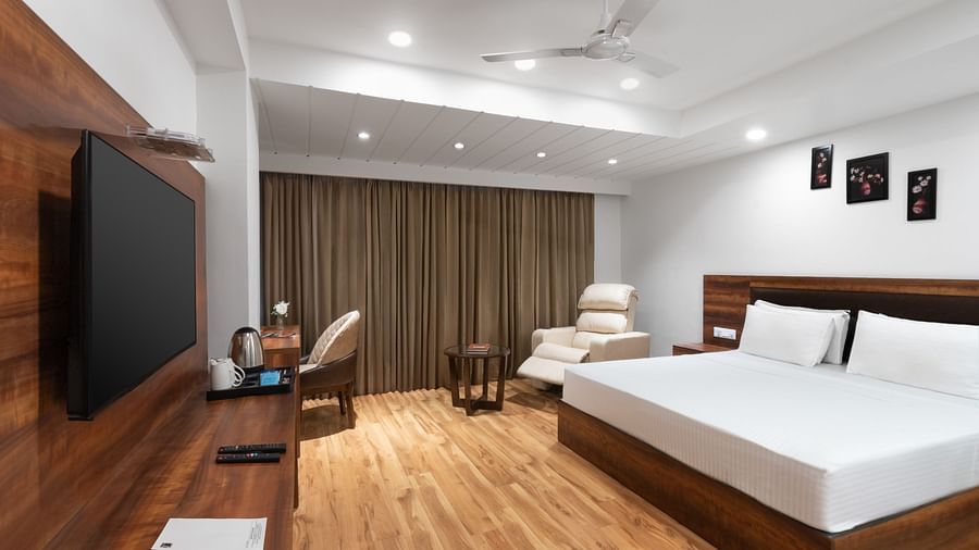 A spacious hotel room with a large bed, wooden flooring, a TV, seating area, and closed curtains.- Clarks Inn Express, Near Jammu Bari Brahmana Railway Station
