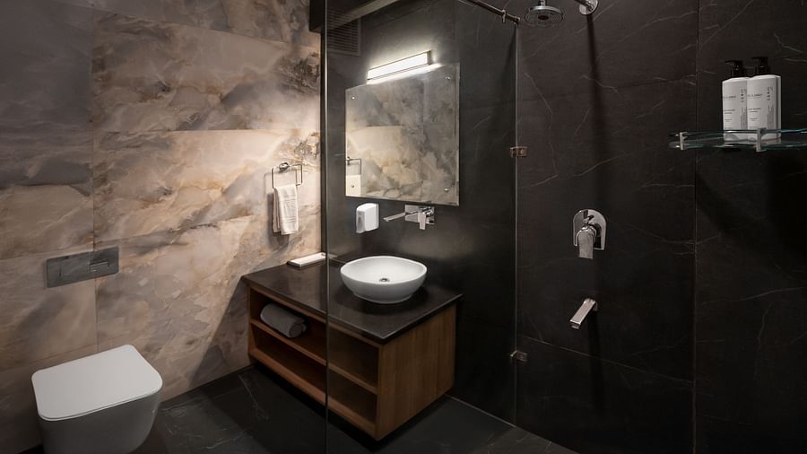  A sleek bathroom with dark marble walls, a black vanity with a round white basin, a wall-mounted toilet, and a glass shower enclosure with a rainfall showerhead. A large illuminated mirror hangs above the sink. - Clarks Inn Express, Near Jammu Bari Brahma