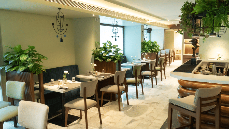 A serene dining area at the Koi Garden of Hotel Bawa International in Vile Parle featuring neatly arranged tables, upholstered chairs, indoor plants, and soft ambient lighting creating a calm and elegant setting.