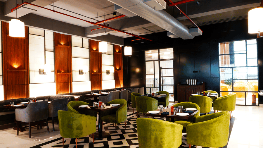 Restaurant at Polo Hotel, Agartala with green velvet tub chairs, dark booth seating, pendant lights and a black-and-white tiled floor under exposed pipes.