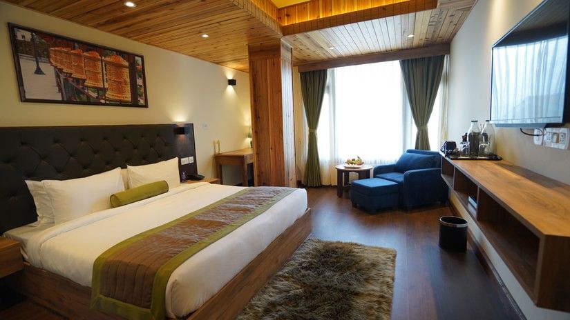 A luxurious room with a wood-panelled ceiling, a king bed with green and brown decor, a lounge chair by the window, and a wall-mounted TV - Mount Hotels