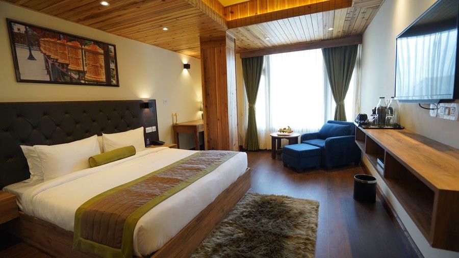 A luxurious room with a wood-panelled ceiling, a king bed with green and brown decor, a lounge chair by the window, and a wall-mounted TV - Mount Hotels