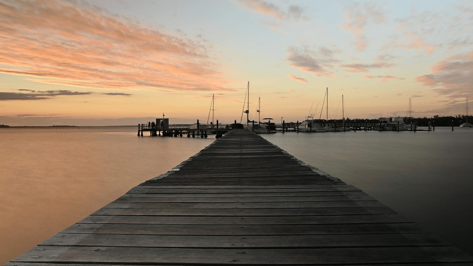 A wooden pier extending into a calm bay, with a beautiful sunset and moored sailboats in the background