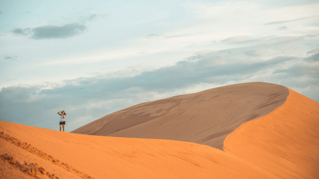 An overview of a desert in mui ne with high red dunes and a woman standing on top in view, one of the best Phan Thiet Tourist Attractions.