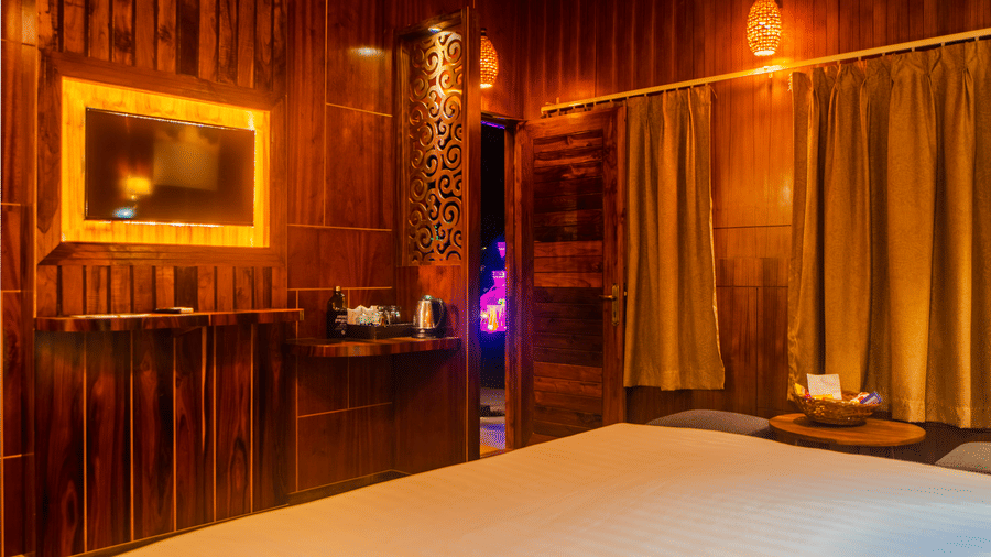 A wooden-themed hotel room featuring warm lighting, a bed with blue accent pillows, and wooden panel walls. The ambient lighting creates a cosy, inviting atmosphere.