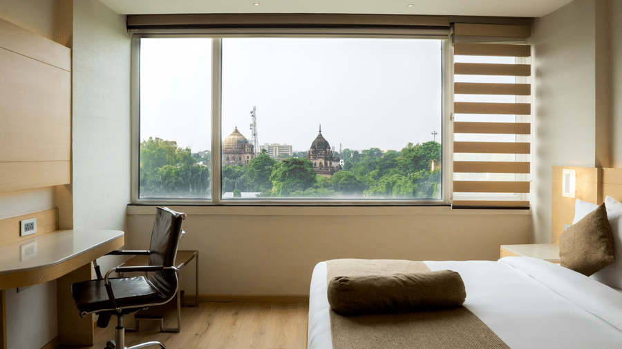 Bedroom of a Suite at Gemini Continental featuring a neatly made bed, and a working area next to a large window overlooking trees and historic monuments outside.