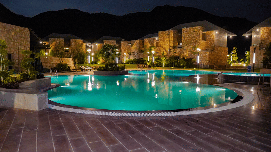 An illuminated swimming pool glows green and reflects the resort buildings at night. The surrounding area is dark with a view of the mountains.