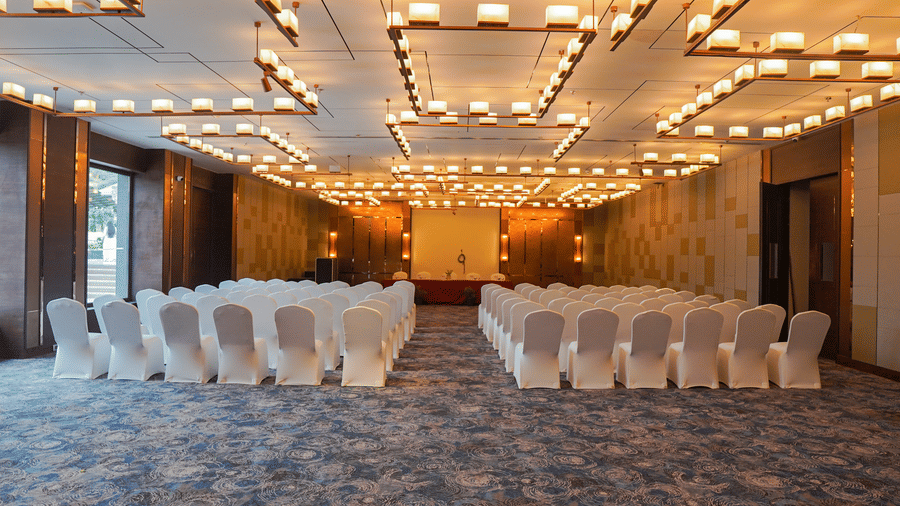 A wide shot of a conference room or ballroom set up with rows of white chairs facing a stage, featuring warm, modern lighting fixtures.