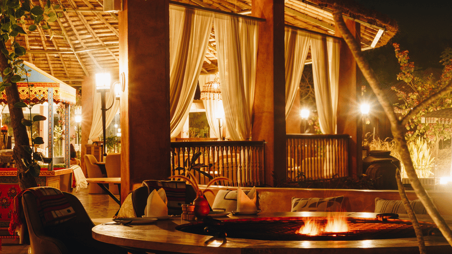 Warmly lit outdoor dining area at Surpura Bagh featuring a central fire pit, wooden seating, draped curtains, and ambient evening lighting.